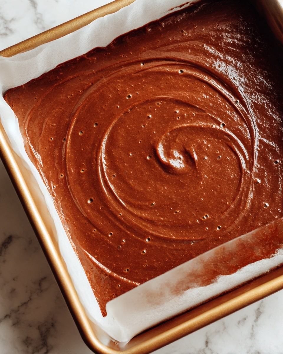 A close-up view of a baking pan filled with smooth, thick chocolate batter. The batter is spread evenly throughout the pan with some gentle swirls and small air bubbles visible on the glossy, rich brown surface. The pan is lined with white parchment paper on the sides, with the metal edges of the pan showing a light golden brown color. The background is a white marbled texture. photo taken with an iphone --ar 4:5 --v 7