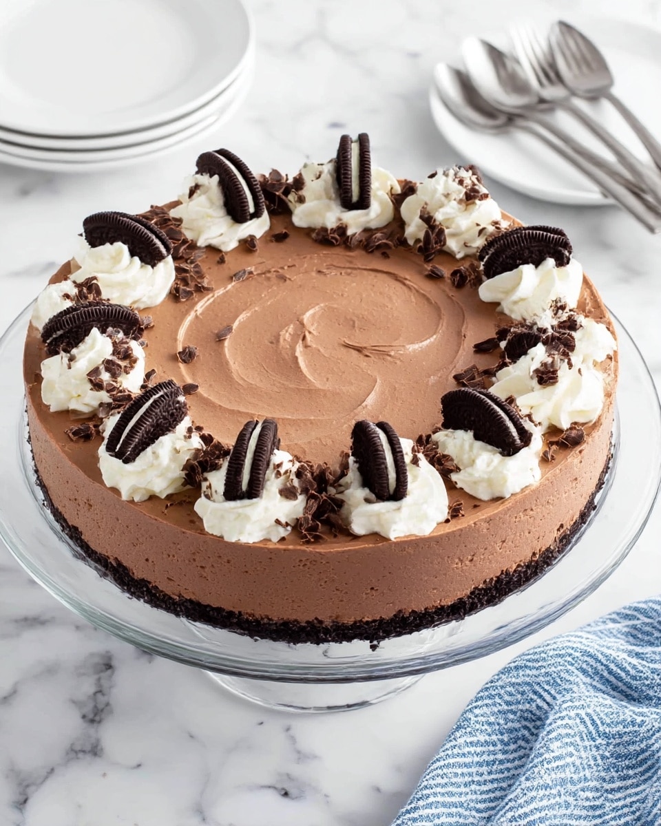 The image shows a round chocolate mousse cake on a clear glass stand placed on a white marbled surface. The cake has a smooth, creamy chocolate mousse layer covering the top, with light swirls creating subtle texture. Around the edge, there are evenly spaced dollops of white whipped cream, each topped with small chocolate curls and half of a sandwich cookie with a dark chocolate exterior and white cream filling. The cake sides are smooth, matching the top layer's color and texture. Nearby, there is a white plate and silver forks resting on the white marbled surface, along with a blue and white striped cloth in the bottom right corner. Photo taken with an iphone --ar 4:5 --v 7