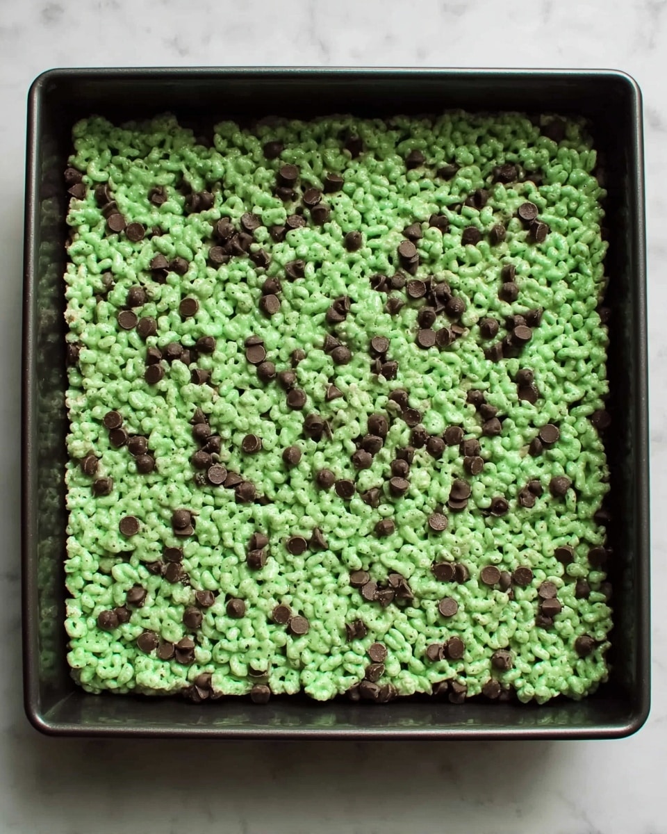A square black baking tray filled with a single thick layer of bright green rice cereal treats mixed evenly with small dark brown chocolate chips, creating a textured and slightly bumpy surface. The treats layer is dense and covers the entire tray, sitting on a white marbled surface. Photo taken with an iphone --ar 4:5 --v 7