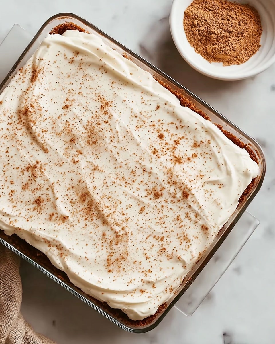 A rectangular glass dish contains a dessert with two distinct layers visible at the edges; the bottom layer is a deep brown color with a crumbly texture, and the top layer is a thick, smooth white cream spread evenly across the entire dessert's surface. The white cream is sprinkled with light brown powder, likely cinnamon or nutmeg, scattered softly over the top. The dish is placed on a white marbled surface, and in the upper right corner, there is a small white bowl filled with the same light brown powder. Photo taken with an iphone --ar 4:5 --v 7