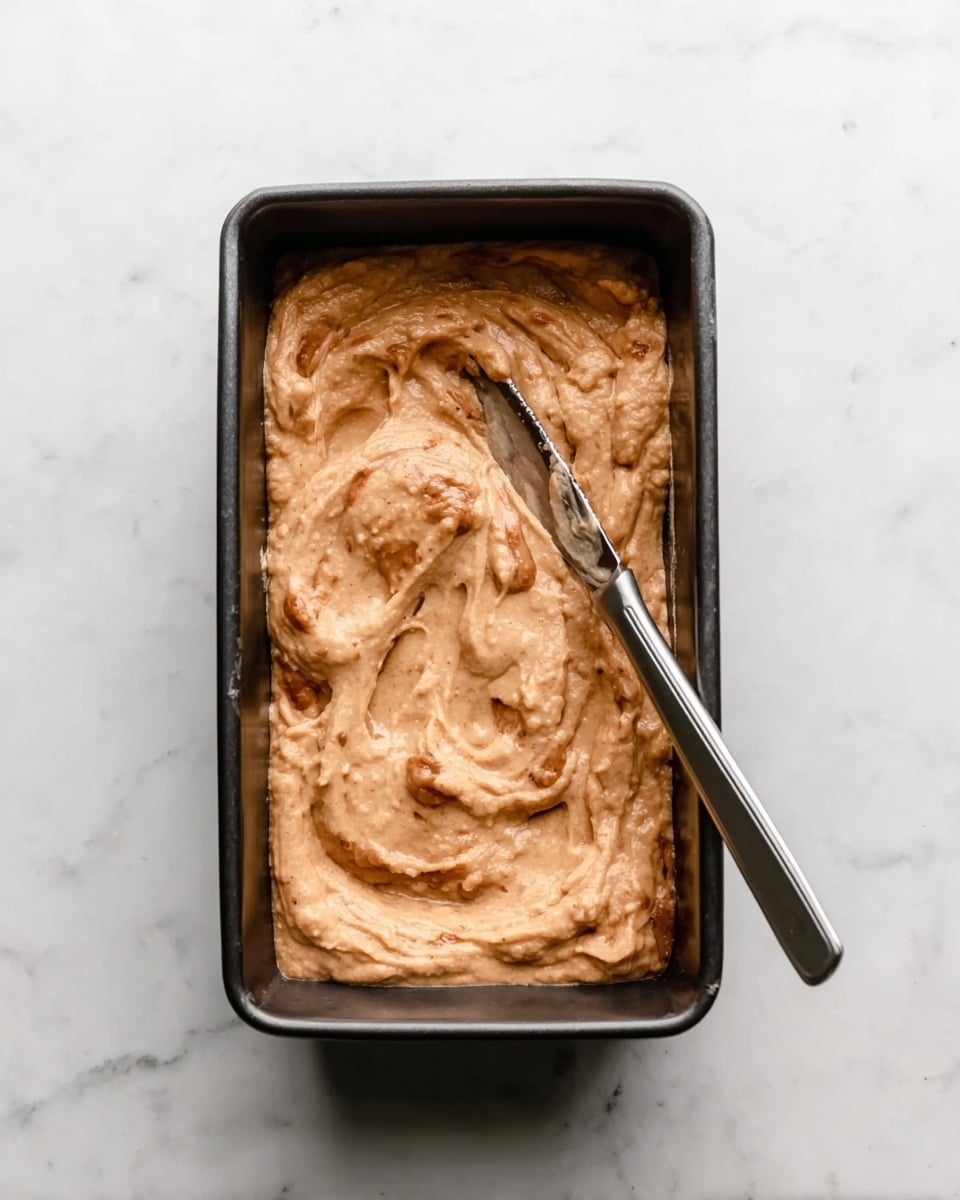 The image shows a rectangular black baking pan filled with a thick, light brown batter that has some darker swirls mixed in. A silver knife is spreading the batter on the surface, leaving soft, wavy texture marks. The pan is placed on a white marbled surface. photo taken with an iphone --ar 4:5 --v 7