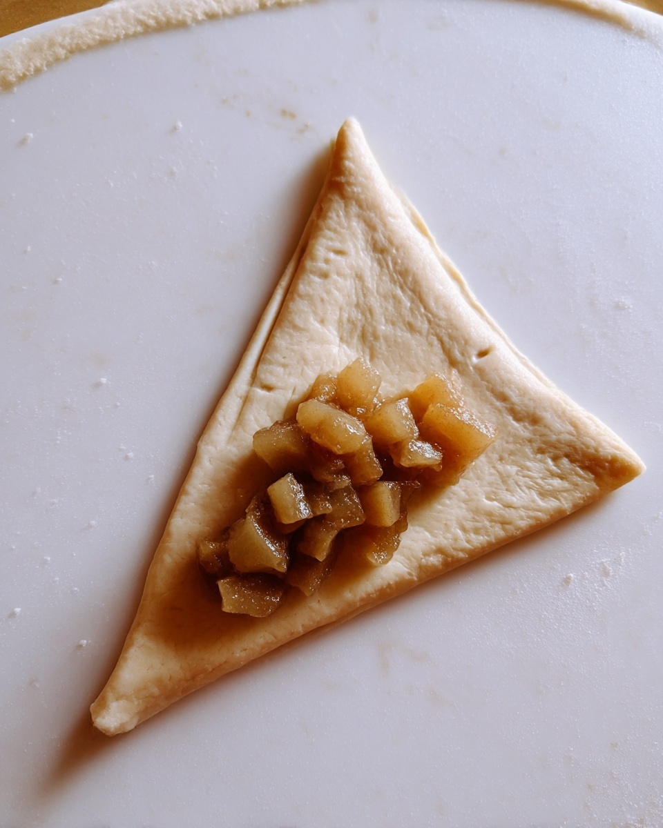A single triangular piece of light beige dough is placed flat on a white marbled surface. Near the base of the triangle, there is a small pile of glossy, brownish diced fruit filling, which looks soft and juicy. The dough has a slightly rough texture with small dented marks along the longer sides. Photo taken with an iphone --ar 4:5 --v 7