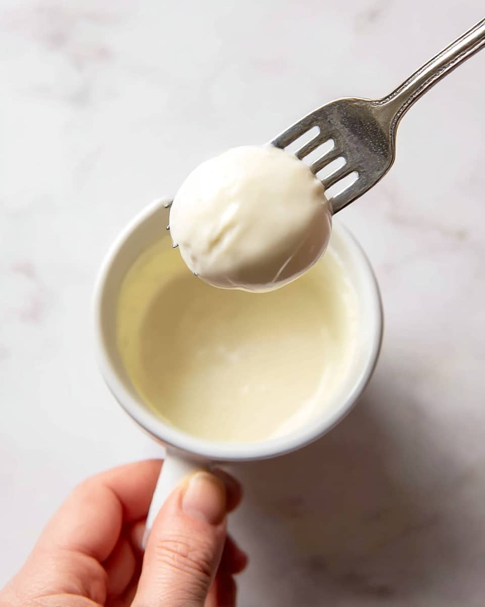 A close-up image shows a white cup filled partially with a smooth, creamy white sauce. Above the cup, a silver fork is holding a round item fully covered in the same creamy white sauce. The background is a white marbled surface with soft lighting, and a woman's hand gently holds the fork at the bottom edge of the image. photo taken with an iphone --ar 4:5 --v 7