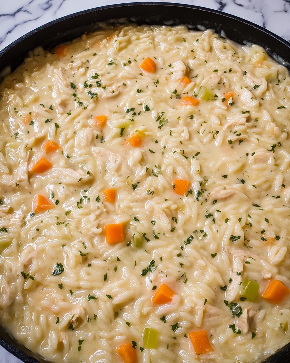 The dish is a creamy one-layer rice and chicken mix cooked in a black pan. The creamy sauce is light beige and thick, holding the rice grains, which are soft and slightly swollen. Mixed into the dish are small pieces of light brown chicken, bright orange carrot cubes, and green celery bits scattered evenly throughout. Tiny green herb flakes are sprinkled on top, adding a hint of color contrast to the creamy texture. The whole content fills the pan closely without overflowing, and the black pan provides a dark frame to the light, creamy food inside. The background is a white marbled surface. Photo taken with an iphone --ar 4:5 --v 7