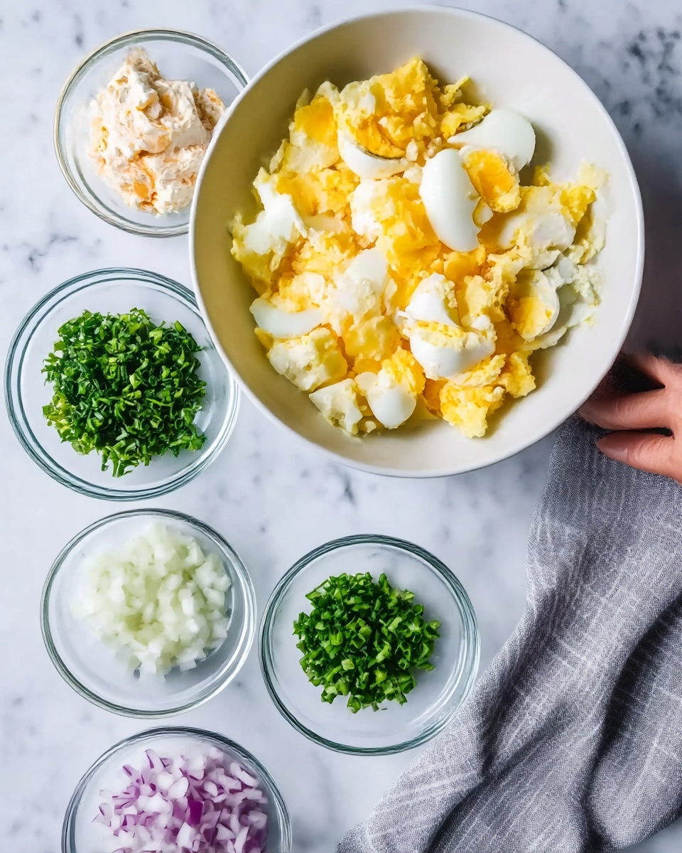 Egg Salad Recipe with the Best Dressing Recipe 4 The image shows a white bowl filled with chopped boiled eggs, mostly yellow yolks mixed with white egg whites. Around this bowl, there are five small clear glass bowls placed on a white marbled surface. One bowl contains a creamy mixture with white and light orange shades, and a little mustard color. Another bowl has finely chopped fresh green herbs, while another has bright chives. One bowl holds finely chopped white onion pieces, and another has small pieces of purple onion. A woman's hand reaches from the right edge of the image, touching a neatly folded gray and white striped cloth next to the egg bowl. Photo taken with an iphone --ar 4:5 --v 7