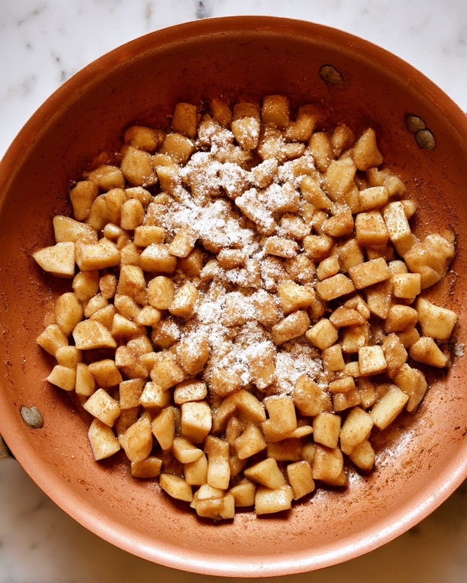 The image shows a copper-colored pan filled with small diced pieces of cooked apples, lightly coated in a brown cinnamon spice. Some white powder is sprinkled on top of the apples, likely flour or cornstarch, which contrasts with the brown and soft texture of the fruit. The apples are spread evenly across the bottom of the pan. The pan is placed on a white marbled surface. photo taken with an iphone --ar 4:5 --v 7