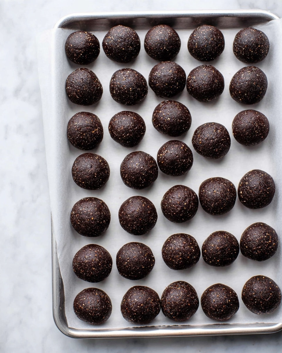 The image shows a silver tray lined with white parchment paper, filled with 25 small, round dark brown balls arranged in a grid pattern. Each ball has a slightly rough texture with tiny light brown specks visible within the dough. The balls are evenly spaced, with some appearing a bit matte while others have a slight shine, hinting at their dense and firm consistency. The background is a white marbled surface, adding a clean and light contrast to the dark balls. photo taken with an iphone --ar 4:5 --v 7