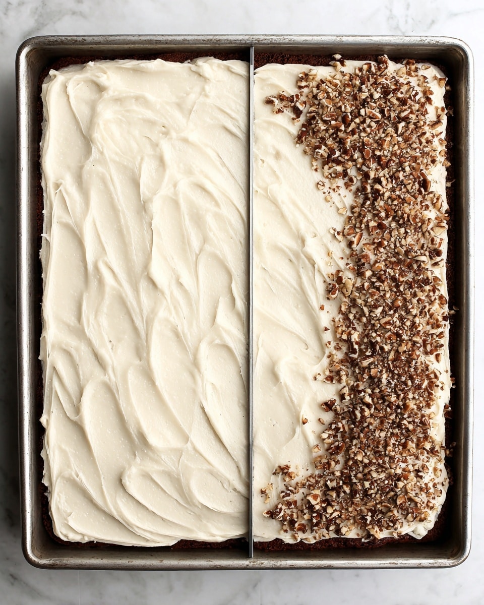 A rectangular cake sits in a metal baking pan with a smooth, thick layer of off-white frosting spread evenly across the top, showing soft swirled textures throughout. On the right side, the frosting is decorated with a generous sprinkling of small, chopped brown nuts that form an uneven border along the edge, giving a crunchy texture contrast to the creamy frosting. The cake base beneath the frosting is a dark brown color, visible slightly around the edges. The pan rests on a white marbled surface photo taken with an iphone --ar 4:5 --v 7