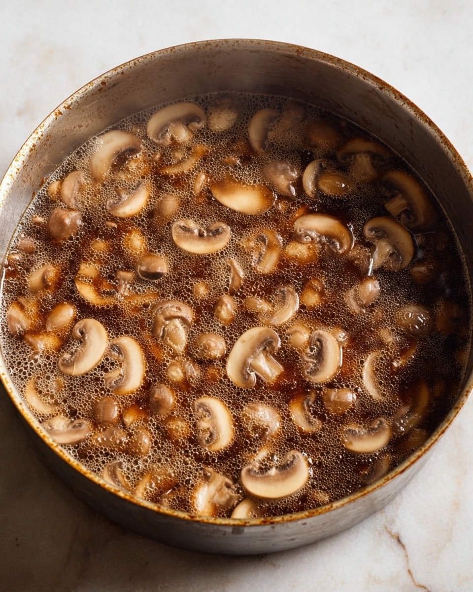 A close-up view of a round metal pan filled with a dark brown liquid, bubbling gently. Inside the liquid are many light brown mushroom slices spread evenly throughout, creating a textured look. The pan has a slightly worn surface, showing signs of use, and it is placed on a white marbled background. Steam rises softly from the hot mixture, adding a warm feeling to the image. photo taken with an iphone --ar 4:5 --v 7