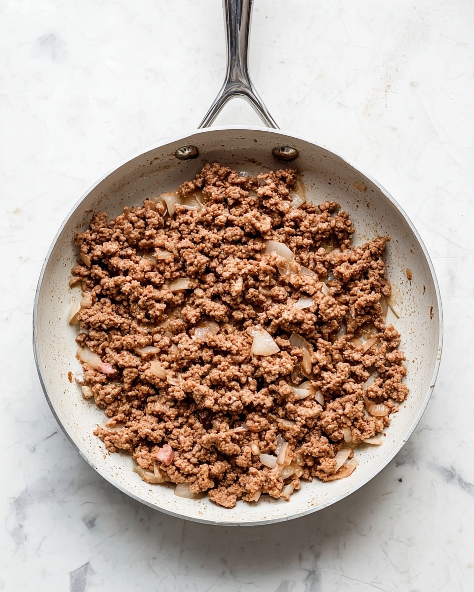 A white pan with a shiny silver handle holds cooked ground meat mixed with small chunks of translucent white onion. The meat is brown and crumbly, spread mostly flat but thick enough to cover the pan's bottom, with some small pink areas visible. The pan is placed on a white marbled surface showing faint grey lines and some scratches. The lighting highlights the texture of the meat and onions, and the image is shot from directly above. photo taken with an iphone --ar 4:5 --v 7