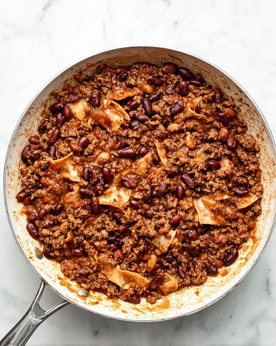 A white pan filled with a rich mix of ground meat, dark red beans, and light brown beans all cooked together in a thick sauce. Scattered throughout are torn pieces of soft, folded light yellow tortilla, blending with the meat and beans. The pan is set on a white marbled surface, and the edges of the pan show slight stains from the cooking sauce. The dish looks hearty and well-mixed, with a warm brown color dominating the scene. photo taken with an iphone --ar 4:5 --v 7