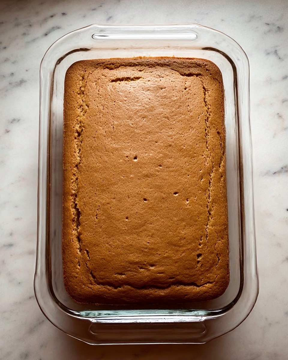 A rectangular, light brown cake with a smooth top surface rests inside a clear glass baking dish with handles on each side. The cake fills the dish nearly to the edges, showing a uniform texture with slight cracks and bubbles on the top. The glass dish is set on a white marbled surface that contrasts softly with the warm tones of the cake. photo taken with an iphone --ar 4:5 --v 7