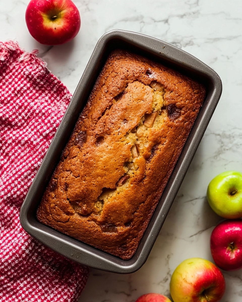 A freshly baked rectangular apple cake with a golden-brown crust, slightly cracked on top showing a bit of the soft texture inside. The cake is still in a dark gray baking pan, placed on a white marbled surface. Around the pan, there are red and green apples and a red and white checkered cloth tucked in the upper left corner, adding a cozy feel. The cake’s surface has uneven browning and close-up details of apple pieces within the cake blend with the brown color. Photo taken with an iphone --ar 4:5 --v 7