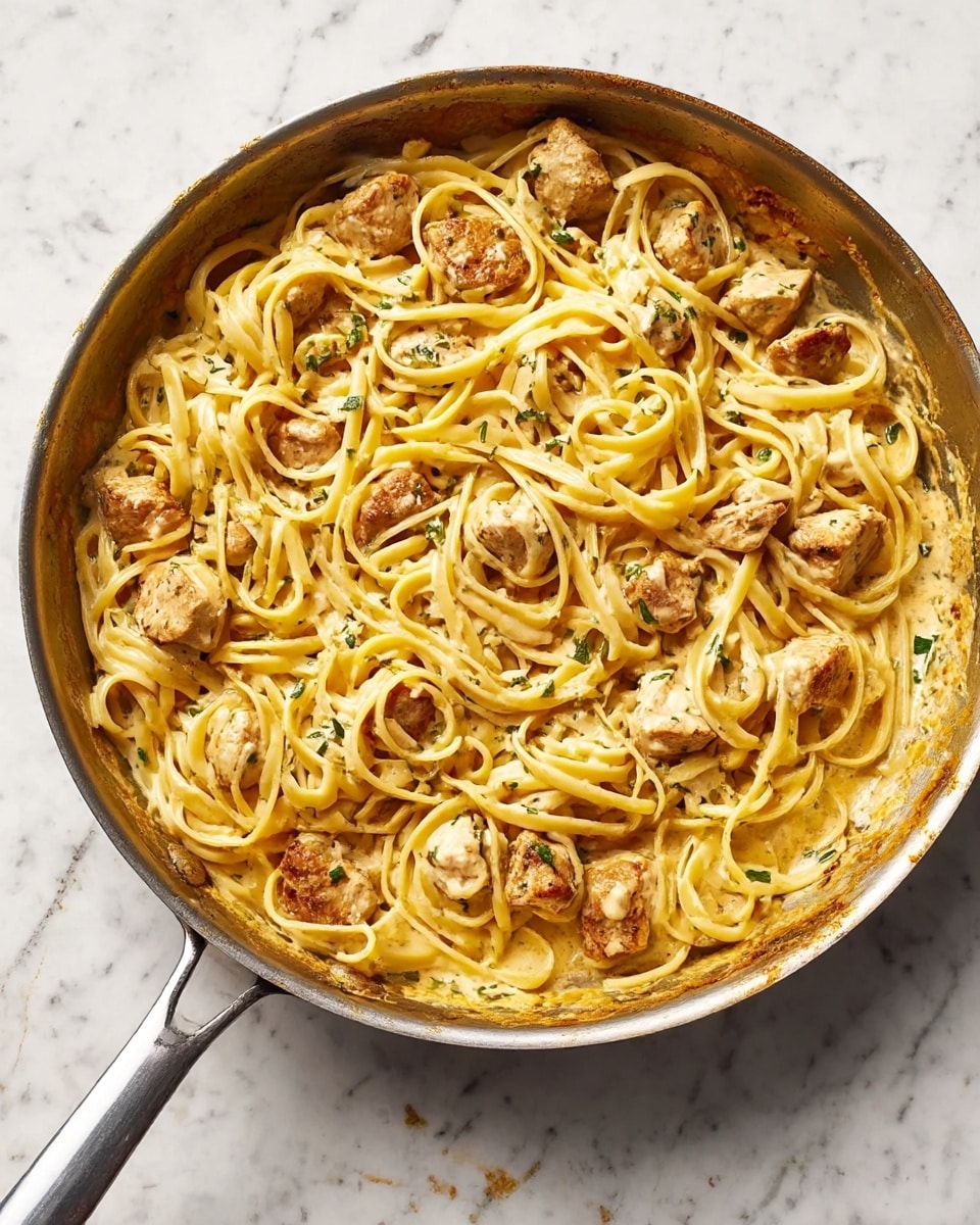 A cooking pan filled with creamy yellow pasta intertwined with chunks of browned chicken pieces, speckled with small green herbs. The pasta looks soft and coated evenly with a light sauce that has a slightly textured surface, while the chicken pieces have a tender and cooked look with a slight golden touch. The pan rests on a white marbled surface, showing some sauce residue around the edges. Photo taken with an iphone --ar 4:5 --v 7