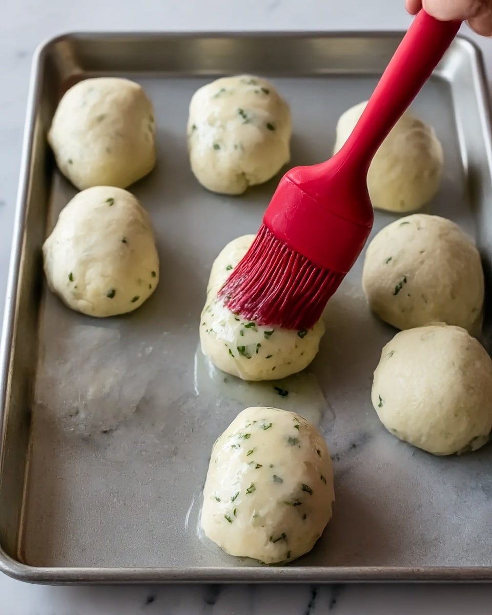 The image shows six small dough rolls arranged on a metal baking tray placed on a white marbled surface. The dough is pale white with some green herb specks mixed in. A woman's hand holds a red silicone brush that is applying a shiny glaze with visible herbs over one dough roll in the front center. The rolls have a smooth, soft texture and are spaced evenly on the tray. photo taken with an iphone --ar 4:5 --v 7