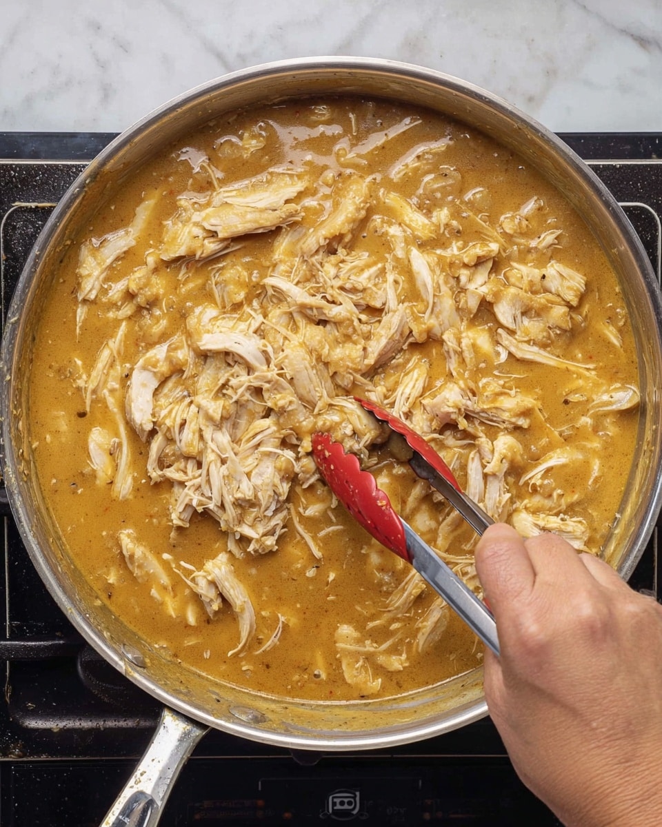 A large silver pan filled with a thick golden brown sauce that covers most of the base, with shredded white chicken pieces mixed into the sauce unevenly across the surface, showing some light browning on the chicken. A woman’s hand holds red-tipped metal tongs stirring the mix from the right side of the pan, which sits on a black stovetop. The sauce has a smooth and slightly shiny texture, and the chicken pieces vary in size scattered throughout the sauce. The scene is set against a white marbled surface. photo taken with an iphone --ar 4:5 --v 7