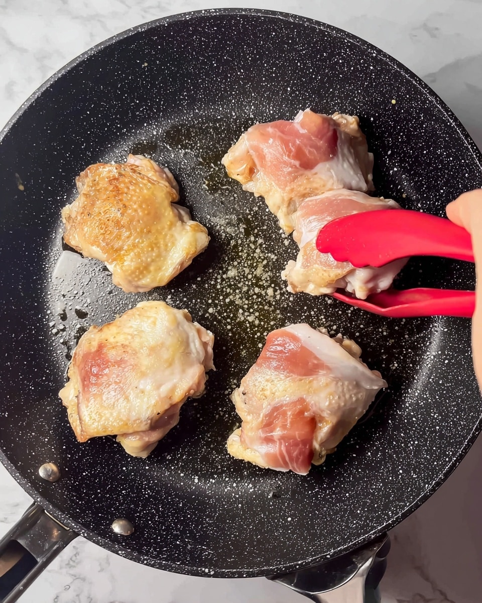A close-up image showing four pieces of chicken thighs being cooked in a black non-stick frying pan with white speckles. Two pieces are golden brown on top, showing they are partially cooked, while the other two pieces are raw with pink skin and white fat visible. A woman's hand with red silicone tongs is holding one of the raw chicken pieces, placing it in the pan. The background is a white marbled texture. photo taken with an iphone --ar 4:5 --v 7