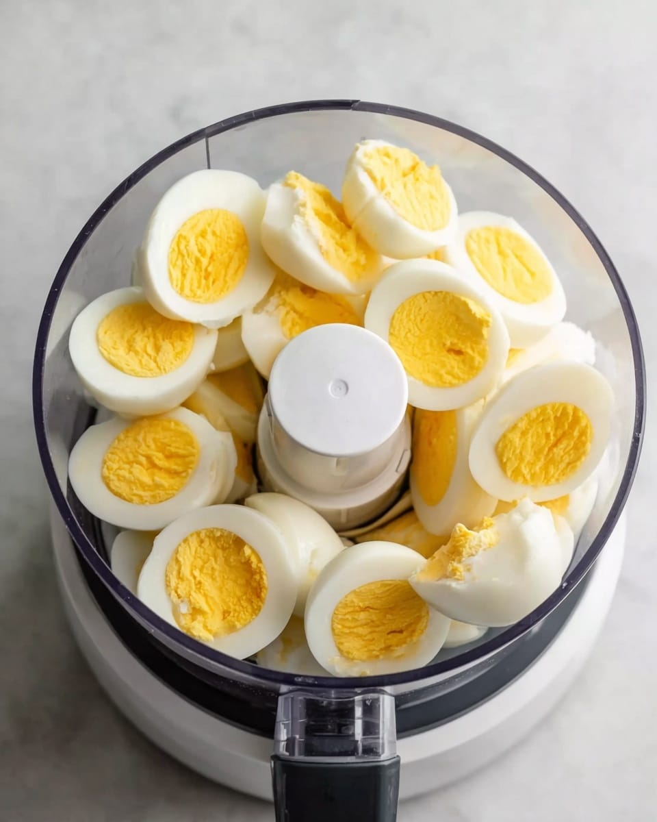 The image shows a clear food processor bowl filled with peeled hard-boiled eggs cut in half, revealing bright yellow yolks surrounded by smooth white egg whites. The eggs are arranged around the central white blade inside the bowl. The food processor sits on a white marbled surface. A woman's hand is not visible in the image but could be implied as nearby. photo taken with an iphone --ar 4:5 --v 7