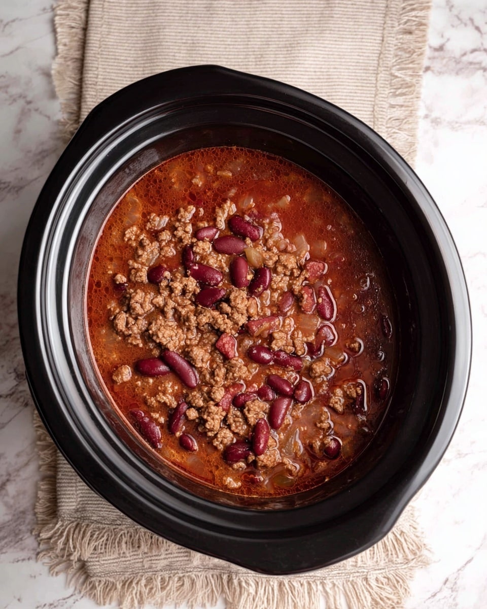 The image shows a black slow cooker filled with a stew-like dish. The stew has three main layers visible: a reddish-brown broth base, scattered dark red kidney beans, and crumbled browned ground meat mixed with small bits of cooked white onion. The mixture looks thick and hearty, with a few bubbles of oil on the surface. The slow cooker sits on a beige cloth with fringe edges, which rests on a white marbled surface. photo taken with an iphone --ar 4:5 --v 7
