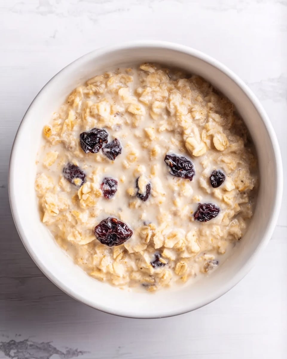 A white bowl filled with creamy oatmeal showing soft, beige oats mixed in a milky liquid. Scattered inside the oatmeal are several dark purple dried berries, adding small dark spots of color. The bowl sits on a white marbled surface, creating a clean, bright background. The texture of the oatmeal looks smooth with some small clumps of oats clearly visible. The image is lit well from above, showing the creamy texture and colors clearly. photo taken with an iphone --ar 4:5 --v 7