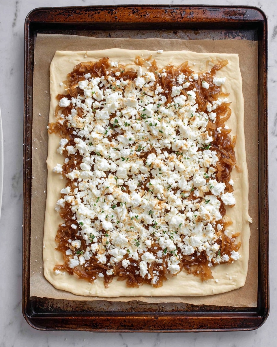A square piece of light beige dough lays flat on a baking tray lined with parchment paper. On top of the dough is a thick, uneven layer of brown caramelized onions spread almost to the edges, showing a glossy texture with some darker and lighter parts. Scattered over the onions is a generous layer of crumbled white cheese, loosely spread, with some small green herb sprinkles visible through the cheese. The baking tray has a dark metal rim with signs of use, and the surface under the tray is a white marbled texture. photo taken with an iphone --ar 4:5 --v 7