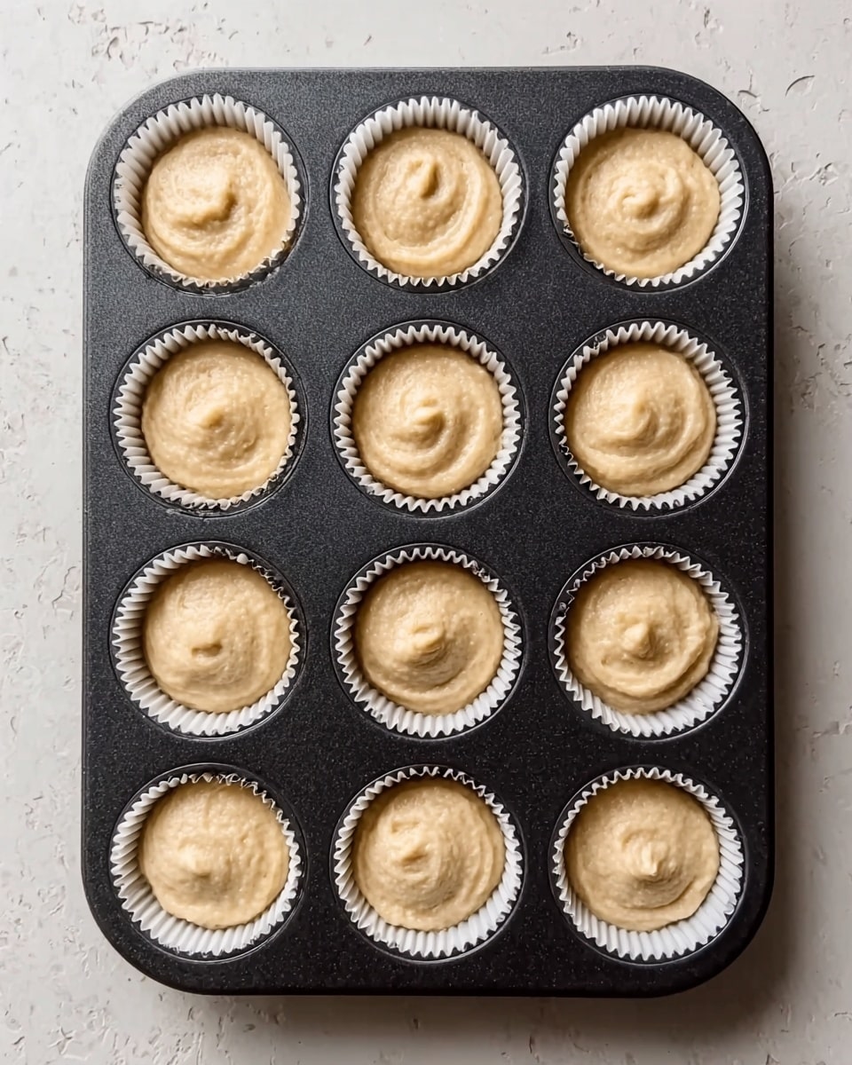 A close-up view of a dark muffin tray with nine white paper liners filled with light beige batter. Each liner is fully filled with batter that looks smooth with some gentle swirls on top. The tray is placed on a white marbled textured surface. There is a slight shadow around the tray edges showing soft natural light. The overall look is neat and ready for baking. photo taken with an iphone --ar 4:5 --v 7