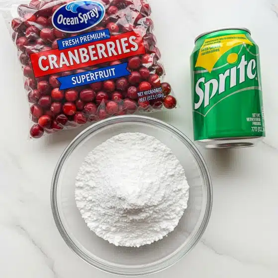 A clear glass bowl filled with a mound of fine white powdered sugar sits on a white marbled surface. Above and slightly to the left of the bowl is a clear plastic bag of bright red cranberries, with a blue and white Ocean Spray label that reads 