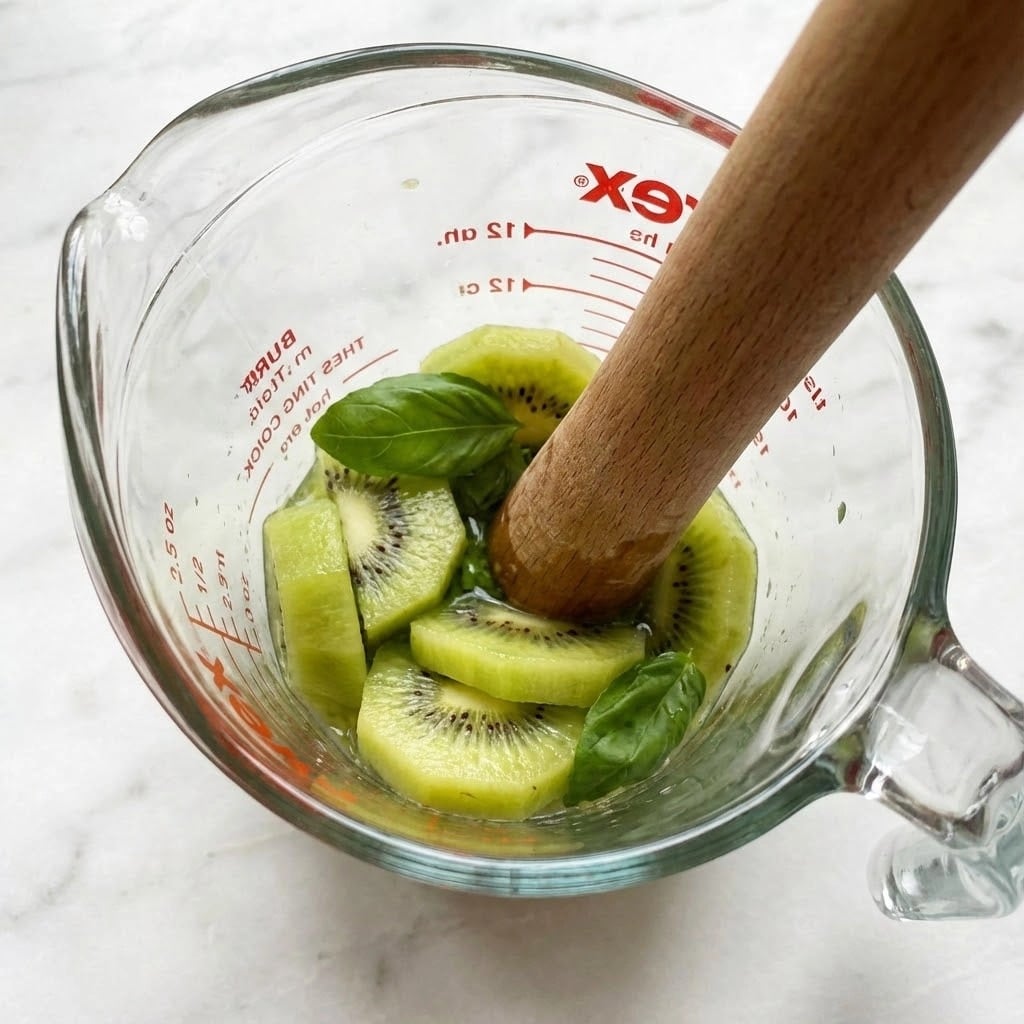 A clear glass measuring cup filled with sliced green kiwi pieces and a few basil leaves, being mashed with a wooden muddler. The kiwi slices show their light green flesh and tiny black seeds, while the muddler is pressing down into the fruit and leaves. The cup has red printed text on the outside, and it is placed on a white marbled surface. photo taken with an iphone --ar 4:5 --v 7