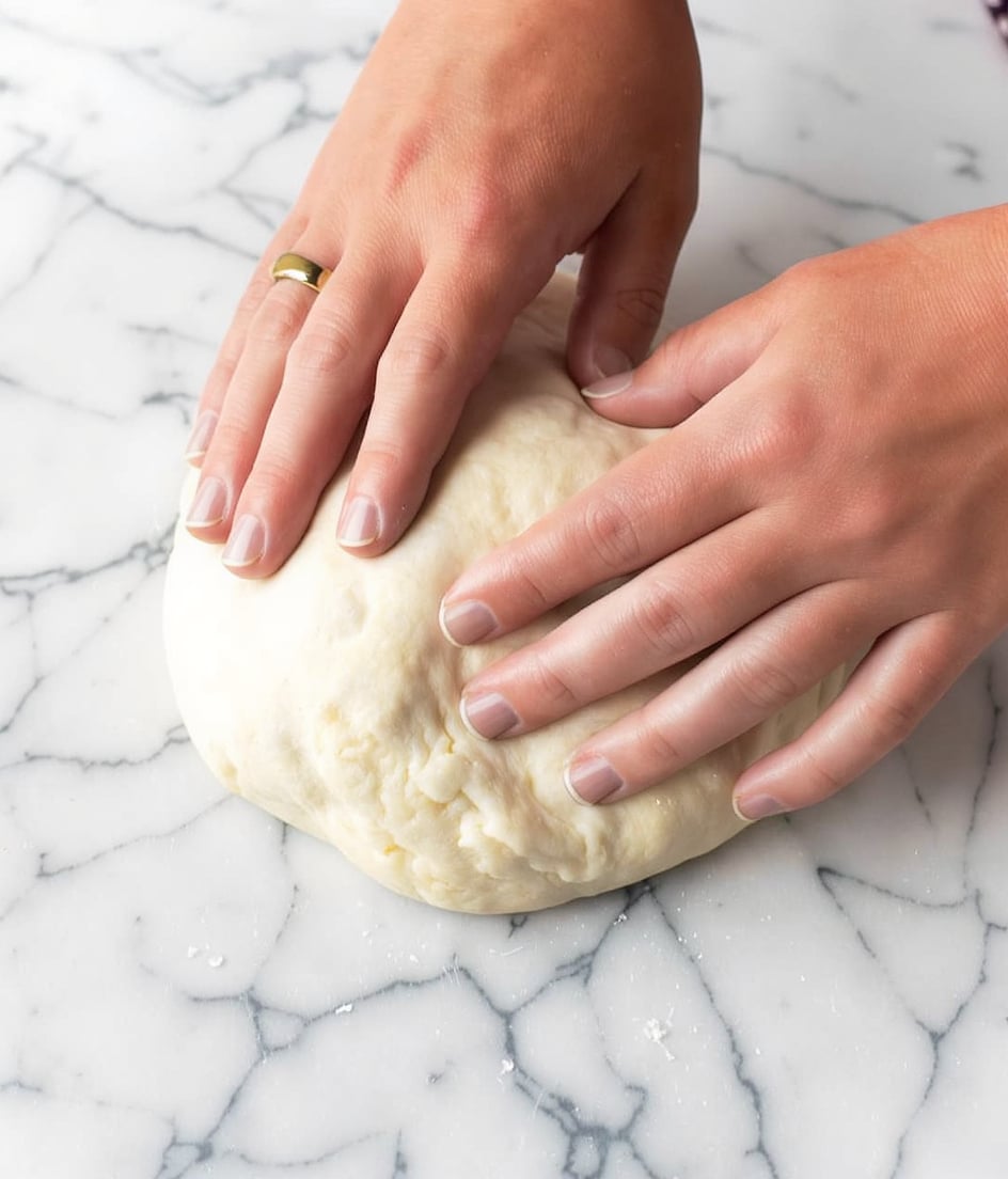 A pair of woman's hands pressing gently on a round, soft dough ball placed on a white marbled surface. The dough is pale beige with a smooth, slightly uneven texture, showing slight stretches where the hands apply pressure. The woman's hands are slightly tanned with a gold ring on the left ring finger. The image focuses closely on the hands and dough, emphasizing the softness and freshness of the dough against the clean white marbled background. Photo taken with an iphone --ar 4:5 --v 7