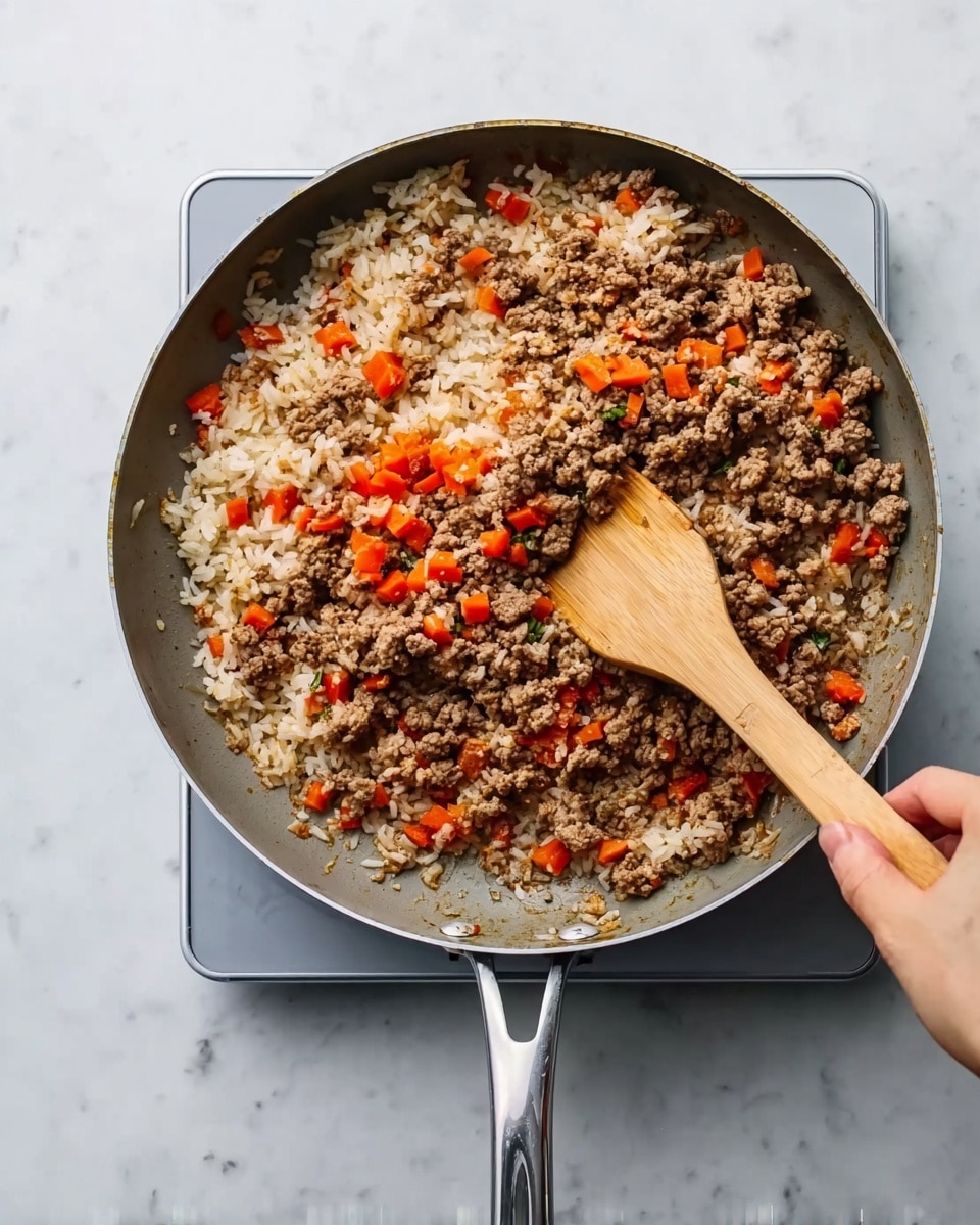 A top view of a deep skillet resting on a white marbled surface with a wooden spatula stirring a mix of cooked ground meat, white rice, and bright orange small diced vegetables. The skillet has a silver handle and contains a fairly even layer of brown textured meat chunks mixed with soft rice grains and scattered small orange pieces throughout. A woman's hand holds the wooden spatula, moving the mixture slightly to the side, exposing some uncooked ingredients below. The scene is simple and clear with the food showing a mix of rough, soft, and small colorful textures. photo taken with an iphone --ar 4:5 --v 7
