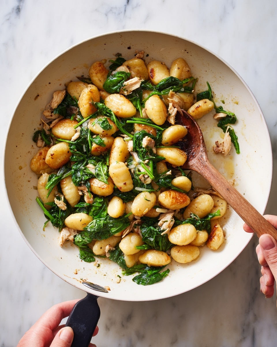 A white pan filled with a cooked mixture of medium-large gnocchi pieces that are golden brown, fresh green spinach leaves scattered unevenly, and small chunks of light-colored chicken. The dish looks warm and slightly shiny from oil or sauce. A woman's hand holds a wooden spoon stirring the food gently inside the pan, while the other hand firmly grips the pan's black handle. The background is a white marble surface. photo taken with an iphone --ar 4:5 --v 7