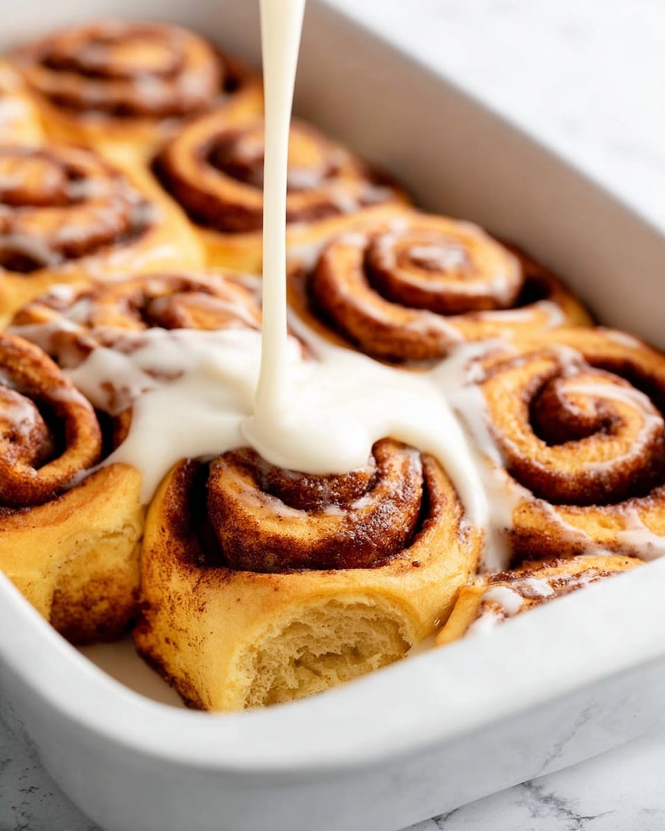 A white baking dish filled with seven cinnamon rolls arranged closely together, each roll showing golden brown layers with a dark brown cinnamon swirl pattern spiraling from the center outward. A thick, creamy white frosting is being poured in a smooth stream onto the cinnamon roll at the front center, spreading gently over its top layer and soft spirals. The background is a white marbled surface, adding a clean, bright contrast to the warm colors of the rolls. Photo taken with an iphone --ar 4:5 --v 7