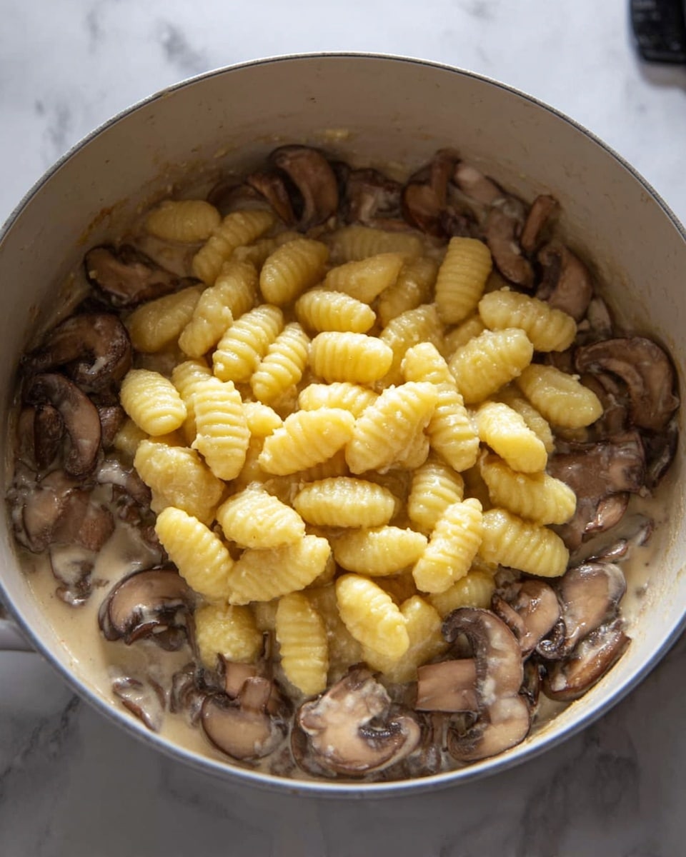 This image shows a cooking pot with two main layers. The bottom layer is filled with sliced brown mushrooms in a light creamy sauce. On top, there is a heap of yellow gnocchi with a ridged and soft texture, piled in the center of the pot. The pot is white, and the background is a white marbled surface. photo taken with an iphone --ar 4:5 --v 7