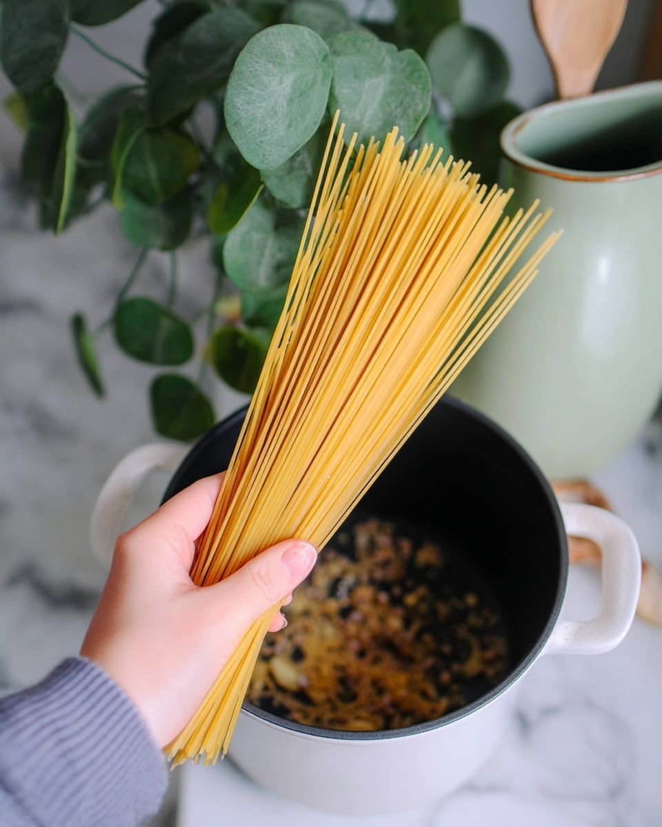A woman's hand holds a bunch of dry, long yellow spaghetti noodles above a black pot with white inside. Inside the pot, there are small pieces of cooked onions or garlic that look brown and soft. The background shows green round leaves of a plant and a light green ceramic jug on a white marbled surface. photo taken with an iphone --ar 4:5 --v 7