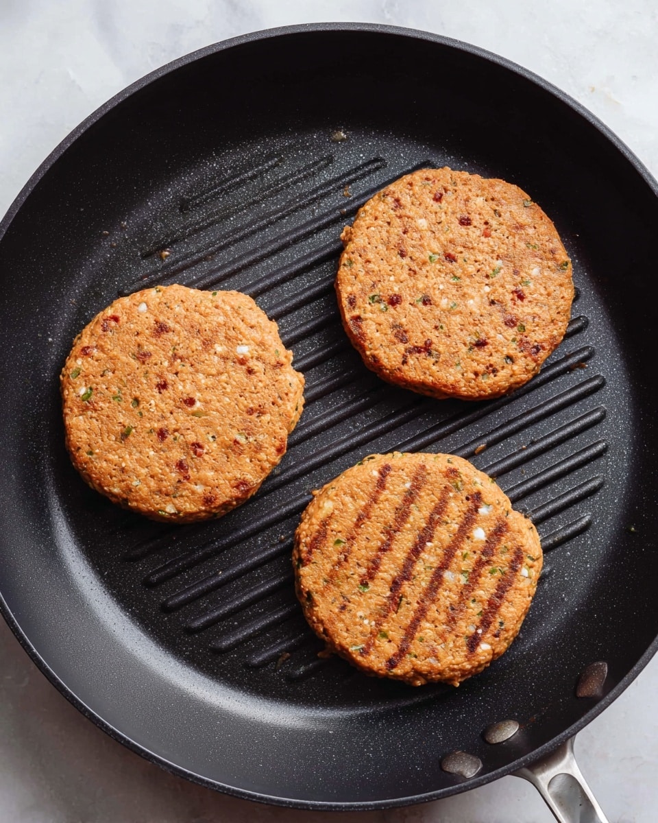 A black round grill pan with ridges contains three orange-brown patties cooking. Each patty is flat and circular, with a slightly rough texture showing small white and green bits inside. The patties are evenly spaced in a triangular arrangement inside the pan. The background is a white marbled surface. Photo taken with an iphone --ar 4:5 --v 7