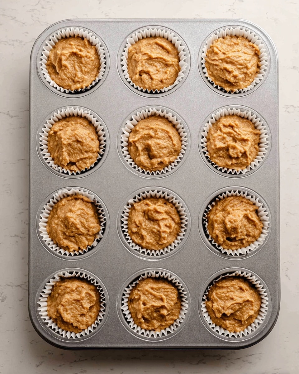 A metal muffin tray holds twelve white paper cupcake liners filled with light brown, thick batter. Each batter portion fills the liners about three-quarters full, showing a rough, uneven texture with small lumps. The tray sits on a white marbled surface, and the top of the batter slightly rises in the center of each liner. The scene shows the muffins ready to bake. photo taken with an iphone --ar 4:5 --v 7
