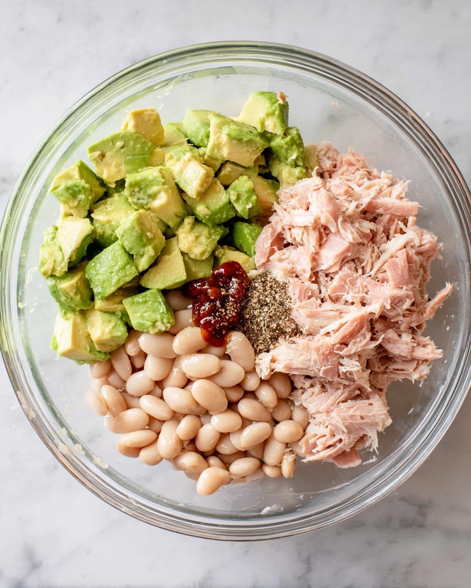 A clear glass bowl on a white marbled surface contains three main layers of ingredients arranged side by side: bright green chunks of avocado on the top left, soft beige white beans on the top right, and shredded light pink tuna at the bottom. On top of the tuna, there are small amounts of ground black pepper and a dash of dark reddish-brown sauce, all ready to be mixed. Photo taken with an iphone --ar 4:5 --v 7