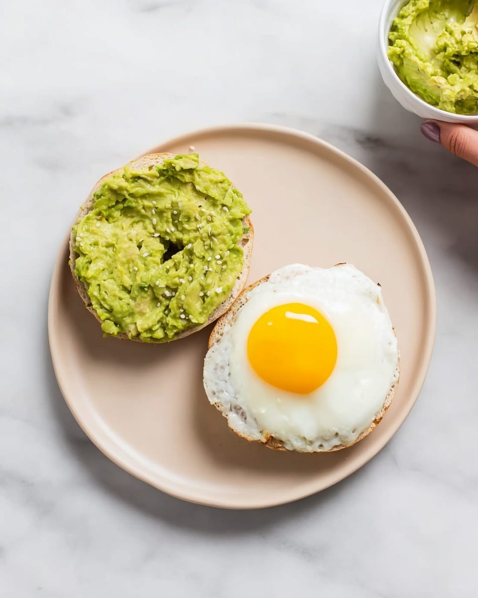 A white plate holds a bagel sliced in half with an avocado spread on the left half, showing a thick, chunky green layer with a rough texture. The right half has a fried egg with a bright yellow yolk sitting on top of the white egg base that looks soft and slightly shiny. The plate is placed on a smooth white marbled surface, and a woman's hand is partially visible near the top right corner holding a bowl with green avocado spread inside. Photo taken with an iphone --ar 4:5 --v 7
