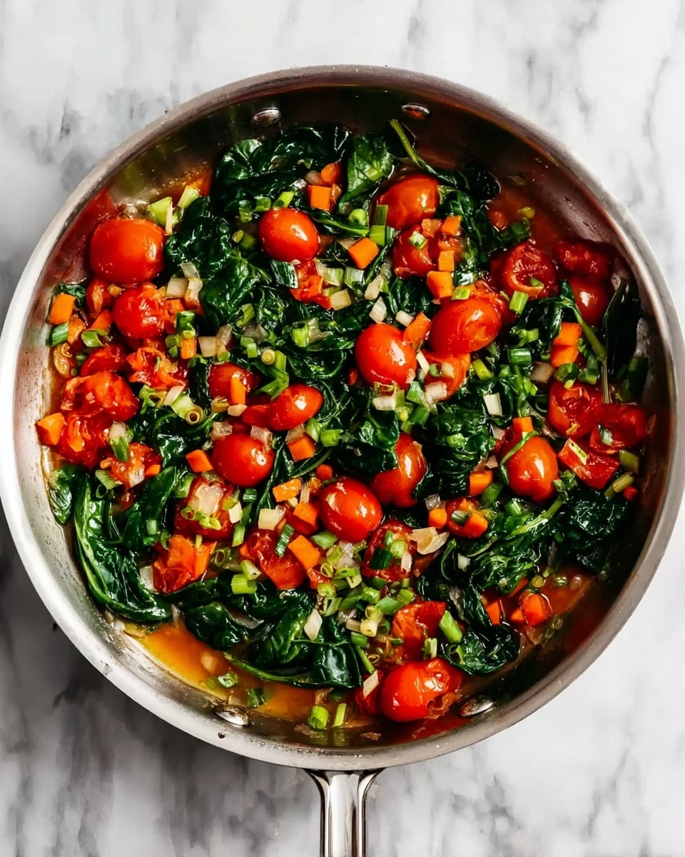 The image shows a shiny silver pan filled with a colorful mix of cooked vegetables on a white marbled surface. Inside the pan, there is a layer of bright red cherry tomatoes, some halved, mixed with small chopped orange carrots scattered throughout. Large dark green spinach leaves add a deep green color, spread evenly across the vegetables. There are also small bits of light green spring onions mixed in, giving texture and color contrast. The pan handle is visible, extending to the right. The light in the photo highlights the freshness and moisture of the vegetables. Photo taken with an iphone --ar 4:5 --v 7