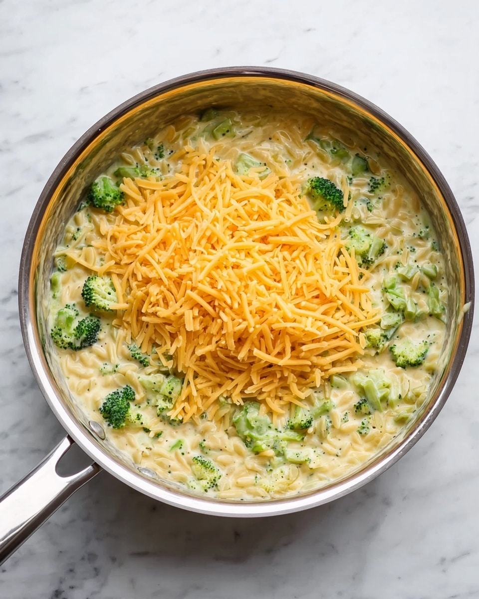 A shiny silver pan sits on a white marbled surface, filled with a creamy mixture of small green broccoli pieces and light yellow pasta. On top of this mixture is a loose pile of bright orange shredded cheese, ready to be stirred in. The pan's handle extends toward the left side of the image, reflecting light. The scene looks bright and clean with soft natural light. photo taken with an iphone --ar 4:5 --v 7