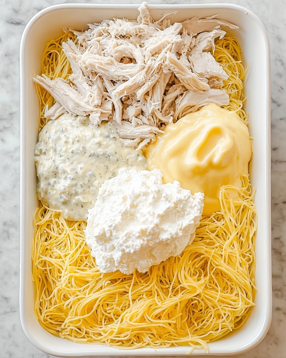 A white rectangular dish holds four main layers spaced apart but not mixed. At the bottom, there is a bed of yellow cooked thin noodles filling the base and sides. Above the noodles, on the top left, is a layer of shredded white chicken with a slightly coarse texture. Next to the chicken on the top right, there is a smooth, shiny dollop of light yellow sauce. At the center left above the noodles, there is a thick, creamy white sauce with small herbs visible inside. In the center bottom above the noodles, a fluffy bright white cream is spread in a round shape. The dish sits on a white marbled textured surface. Photo taken with an iphone --ar 4:5 --v 7