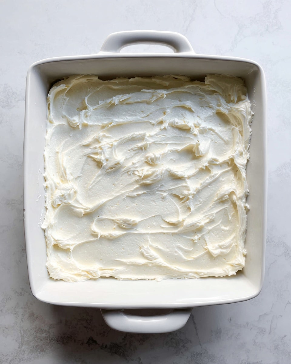 A white square dish with handles is filled with a thick layer of creamy white frosting that is spread unevenly, creating texture with visible swirls and soft peaks. The dish sits on a white marbled surface that adds a clean and elegant backdrop to the simple and smooth frosting layer. photo taken with an iphone --ar 4:5 --v 7