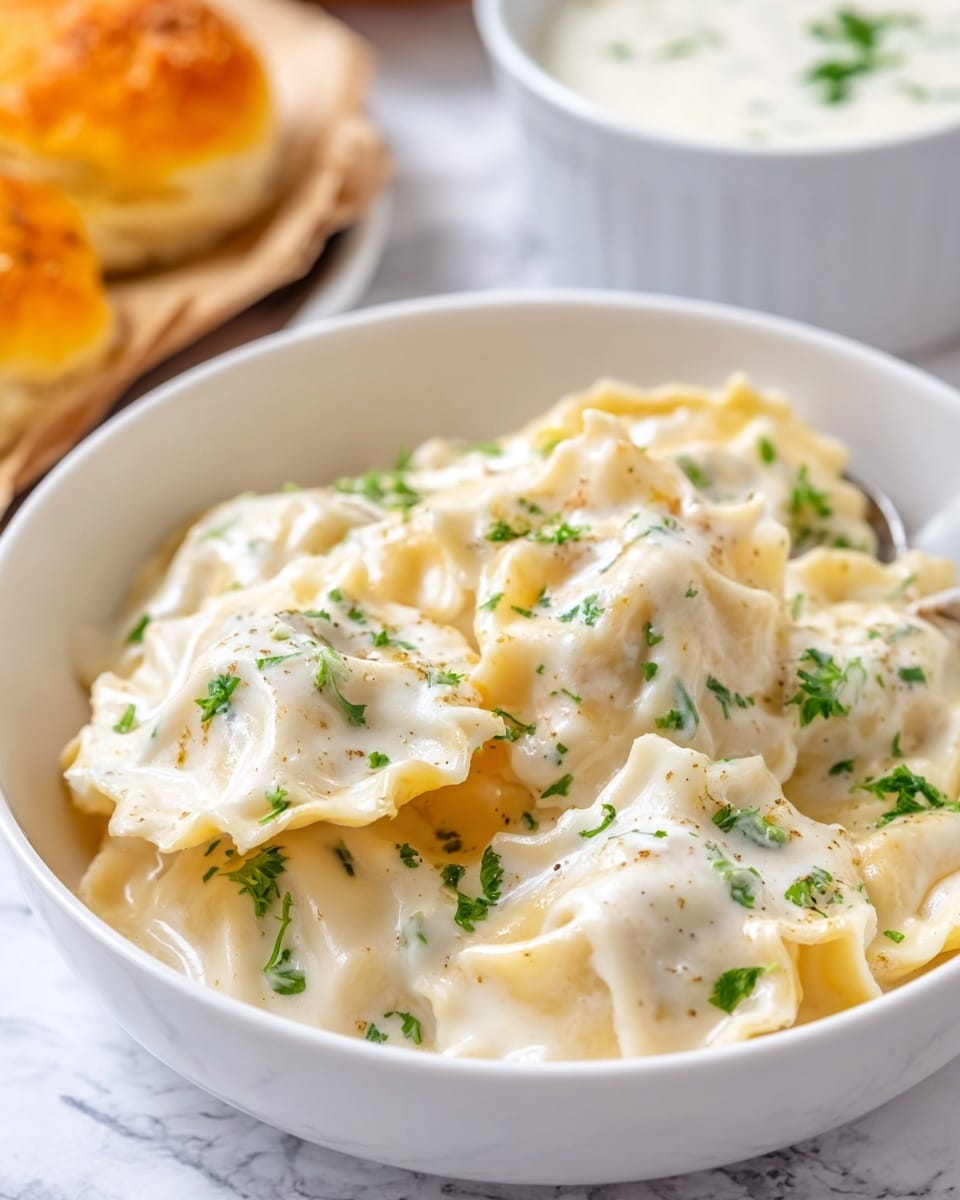A white bowl filled with several pieces of ravioli pasta covered in a creamy white sauce, each piece showing ruffled edges and a soft texture, scattered with small green parsley leaves on top for color contrast. Behind the bowl, there is a soft focus on part of an orange biscuit or roll and another white bowl with white sauce, all set on a white marbled surface. The image captures a warm close-up view that highlights the creamy texture and fresh herbs. Photo taken with an iphone --ar 4:5 --v 7