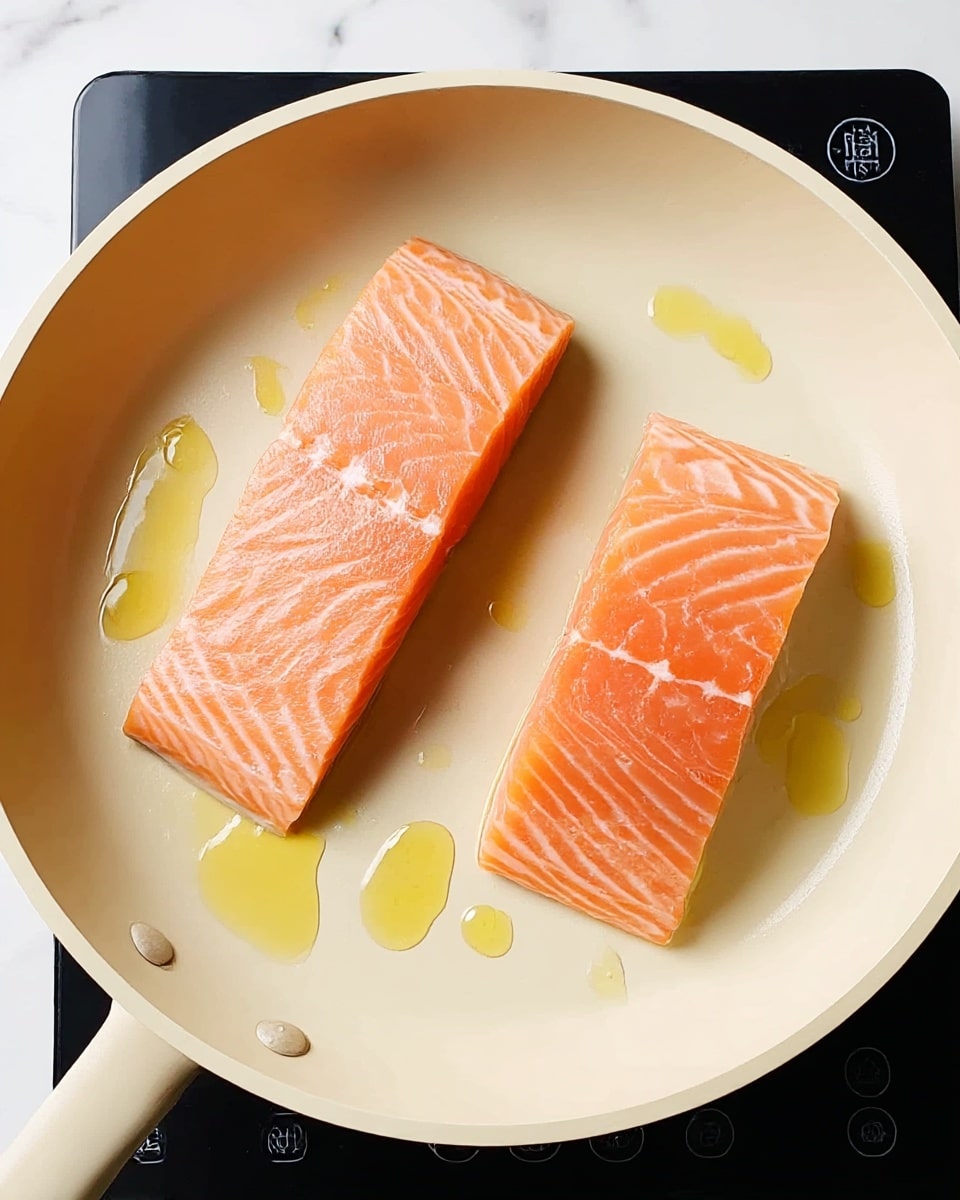 Two raw salmon fillets with a pinkish-orange color and fine white lines are placed flat in a cream-colored frying pan. The fillet on the left is rectangular and smooth, while the one on the right has a slightly uneven shape and a shiny wet texture. Drops of yellow cooking oil are spread around them, creating small shiny puddles. The pan rests on a black stove with control icons visible in the top left corner. The background is a white marbled surface. Photo taken with an iphone --ar 4:5 --v 7