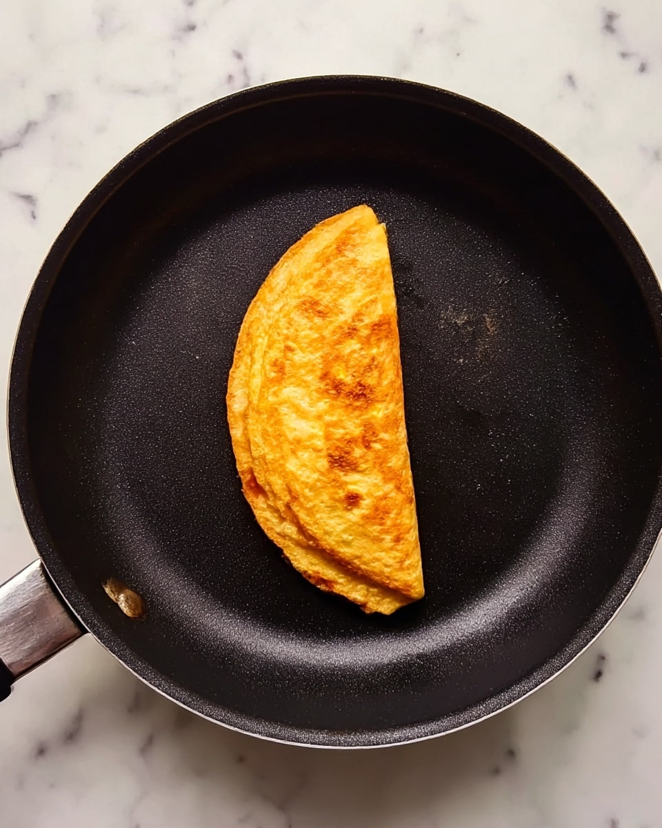 A single folded golden-brown omelette sits in the center of a black non-stick frying pan with a slightly textured surface; the omelette's edges are smooth and slightly crisp, showing a few small darker spots where it cooked more. The pan rests on a white marbled surface with faint light gray veins. The background is simple and clean. photo taken with an iphone --ar 4:5 --v 7