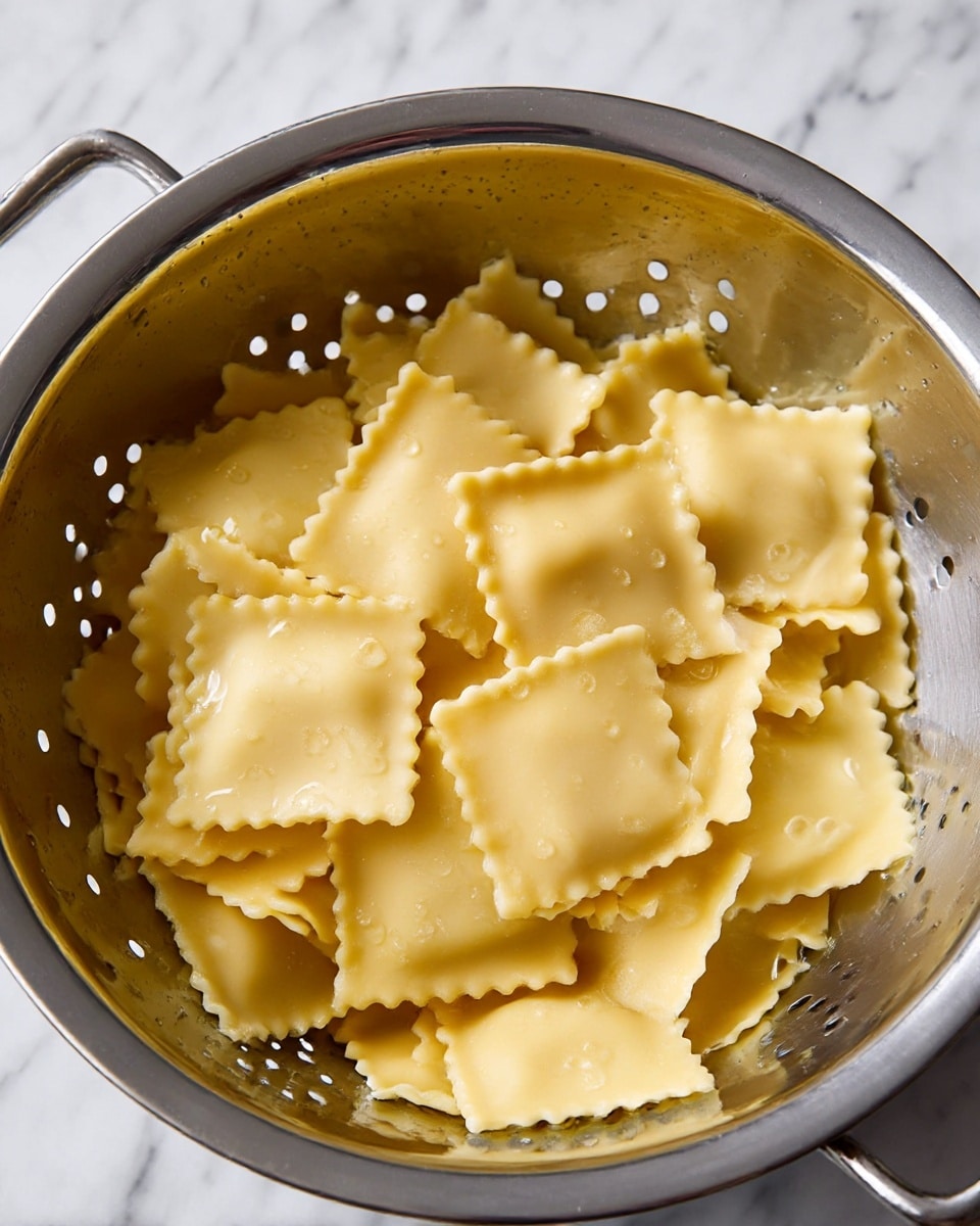 The image shows a silver metal colander filled with cooked ravioli pasta. The ravioli pieces are square with a light yellow color, some having ruffled edges while others have straight edges. The pasta looks soft and slightly wet with small drops of water on and around them. The colander has small holes evenly spaced around it and is set on a surface with a white marbled texture. The lighting reveals the smooth, slightly shiny texture of the pasta photo taken with an iphone --ar 4:5 --v 7