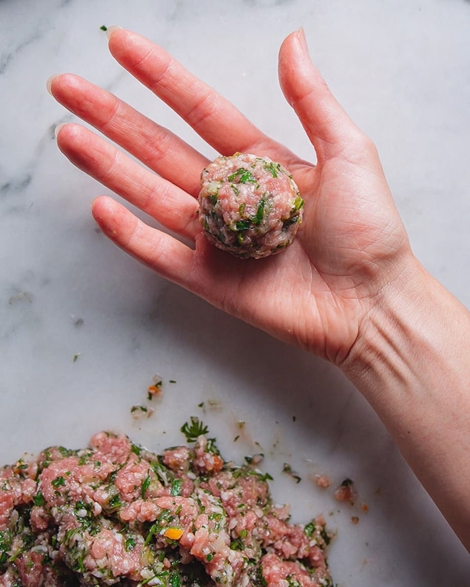 A woman's hand is holding a small ball of raw minced meat mixed with green herbs and small white and orange bits. Below the hand, a larger amount of the same mixture is laid out, showing a rough, soft texture with visible chopped herbs and seasoning. The background is a white marbled surface. photo taken with an iphone --ar 4:5 --v 7