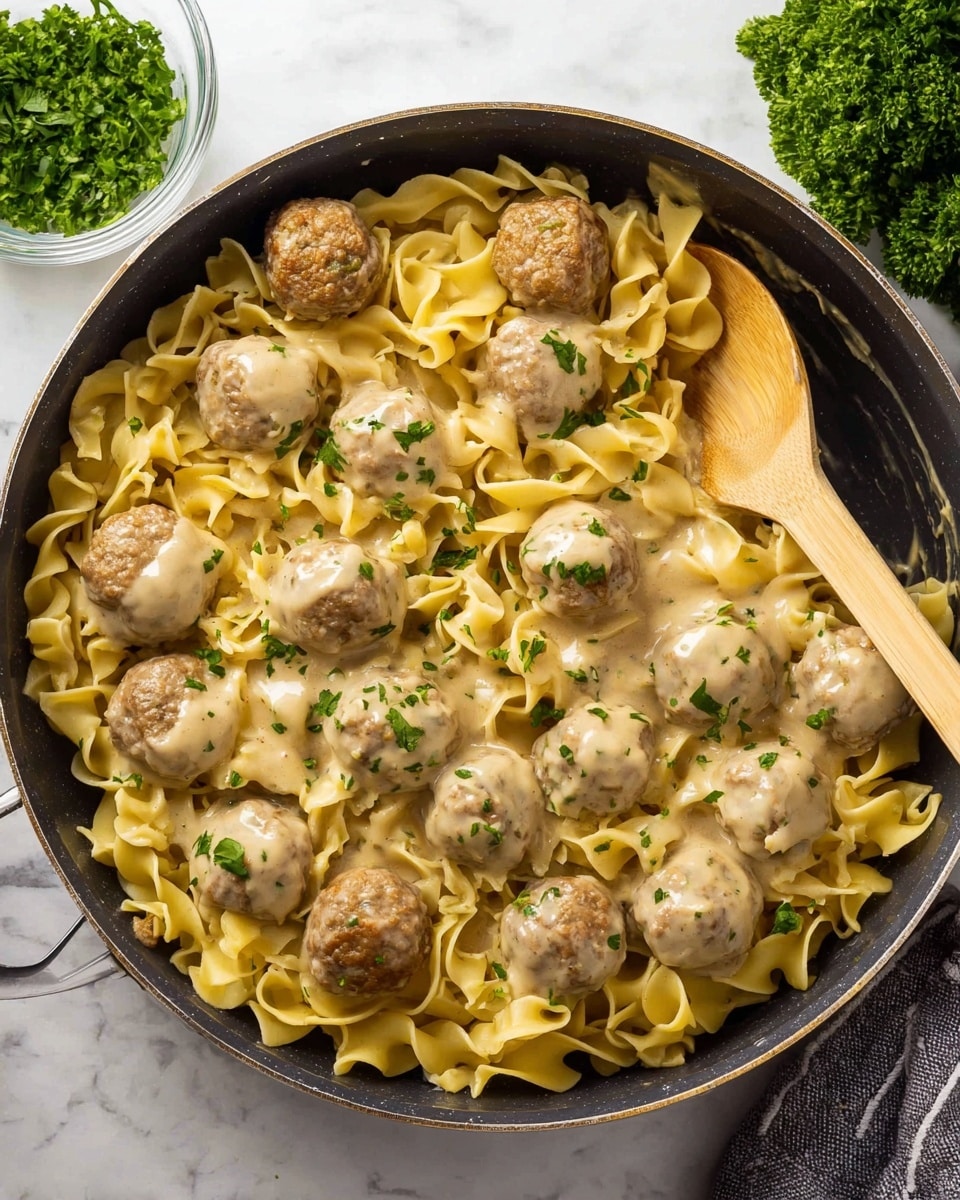 The image shows a frying pan filled with three layers: the bottom layer is wide, curly, yellow pasta with a soft texture, the middle layer consists of round, light brown meatballs evenly spread over the pasta, and the top layer is a creamy beige sauce coating both pasta and meatballs lightly. The dish is garnished with small sprinkles of chopped green herbs. A wooden spoon with a pale handle rests on the right side inside the pan. The pan is placed on a white marbled surface, with a small glass bowl of finely chopped green herbs in the top left corner and fresh parsley visible in the top right. photo taken with an iphone --ar 4:5 --v 7