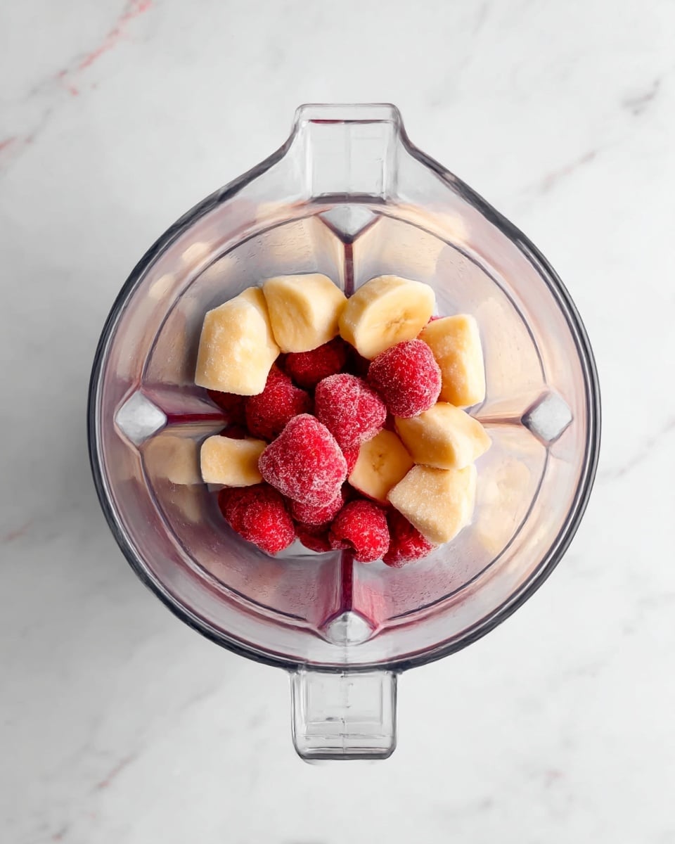The image shows a clear blender container placed on a white marbled surface, containing two layers of frozen fruit. The bottom layer has several chunky slices of pale yellow frozen banana, while the top layer consists of whole frozen strawberries with a bright red color and icy texture. The fruits fill the center of the blender container, with the clear sides and handle visible from above. photo taken with an iphone --ar 4:5 --v 7