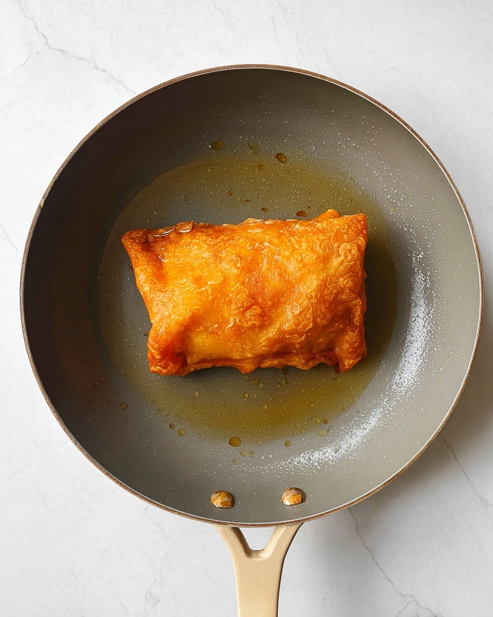 A single piece of fried food with a golden brown, crispy outer layer sits in the center of a gray pan with beige handles. The fried item has a slightly uneven surface with some folds and raised edges, showing texture and crunchiness. The pan has a little bit of oil spread around the fried piece, reflecting light softly. The scene is set against a white marbled surface that gives a clean and bright background. photo taken with an iphone --ar 4:5 --v 7